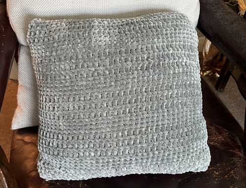 A grey velvety-looking cushion made in stripes of chunky crochet.

The cushion is leaning against a larger cream cushion on a brown chair.

It's about 12 inches / 30cm square. 